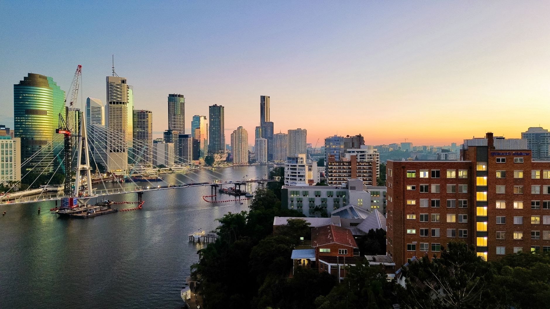 Aerial view of Brisbane's skyline and river at sunset, with a construction crane beside a cable-stayed bridge.