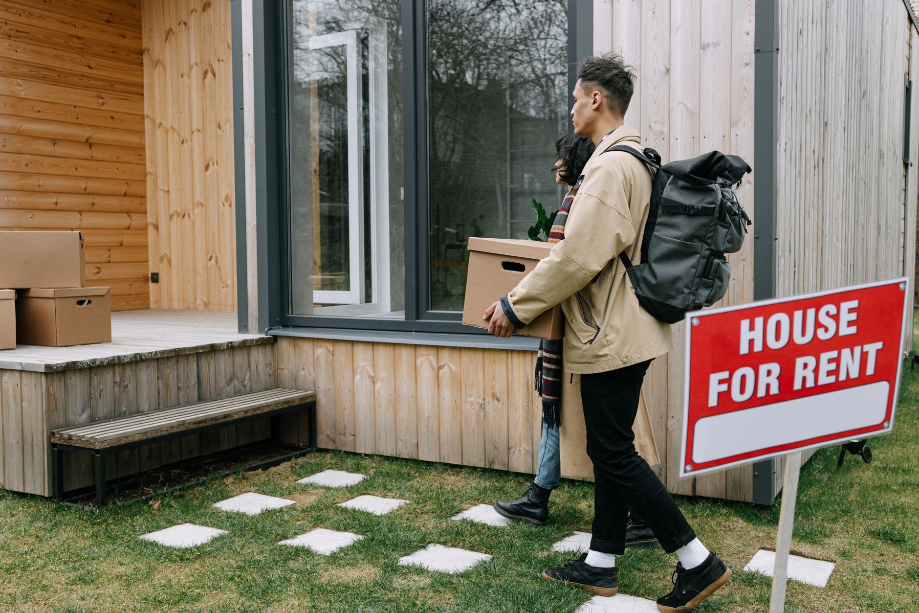A couple carrying boxes and a backpack walk toward a wooden house with a 'House for Rent' sign on the lawn.