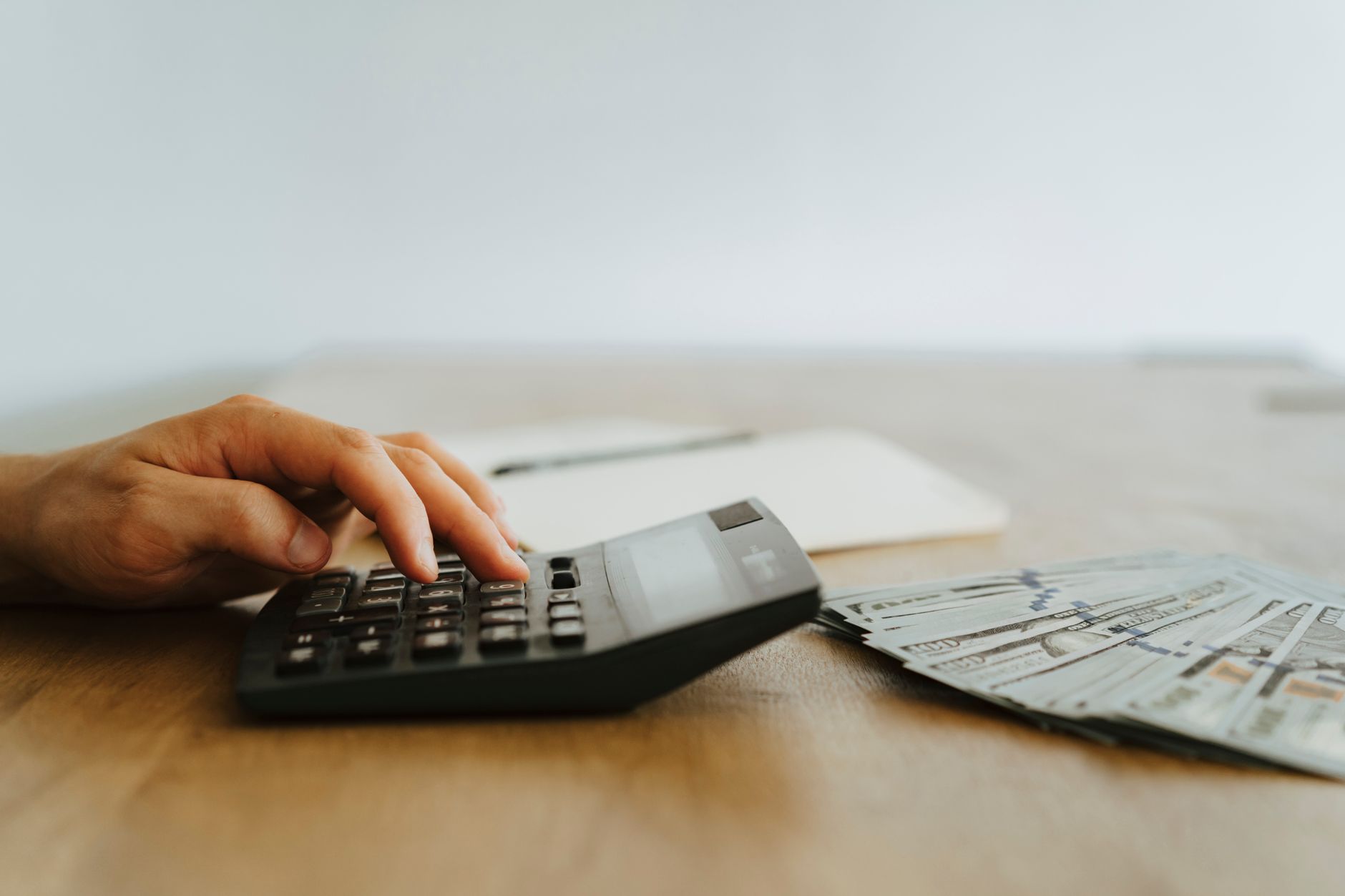A hand presses buttons on a calculator beside a fanned stack of hundred-dollar bills on a wooden desk.